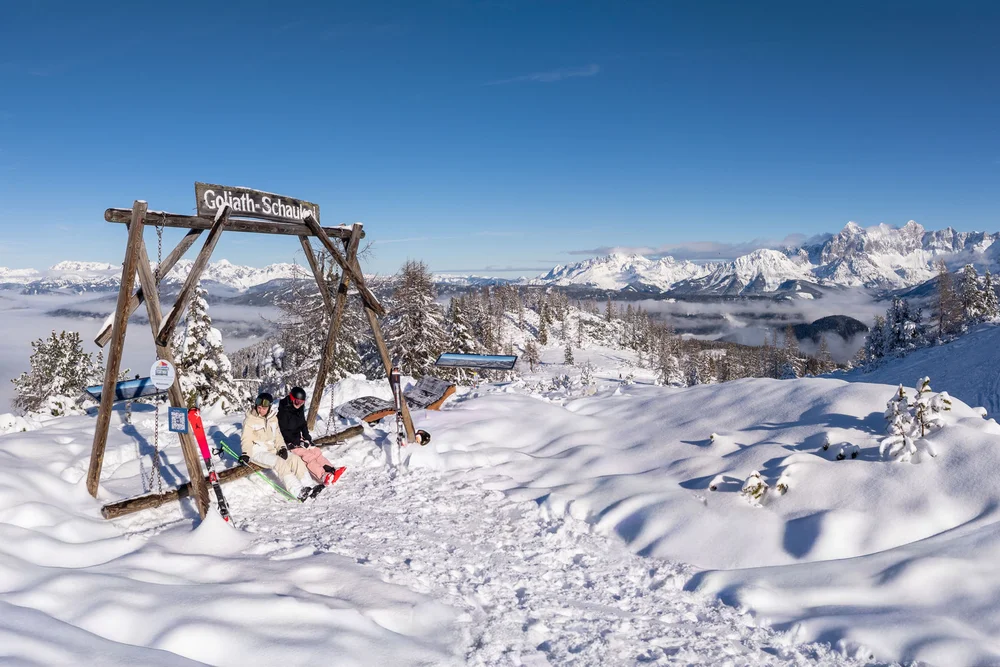 Zwei Personen auf der Goliath-Schaukel vor einer verschneiten Bergkulisse