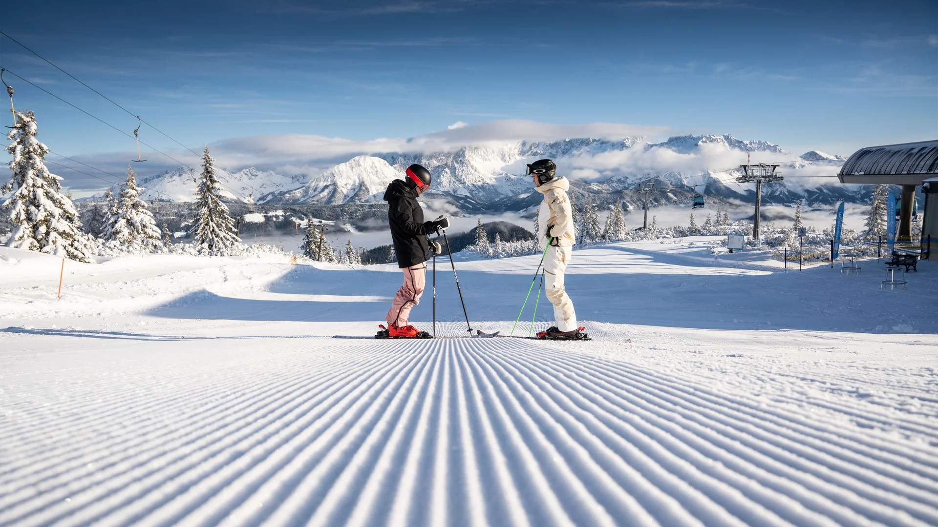 Zwei Skifahrer auf einer präparierten Piste vor einer verschneiten Bergkulisse
