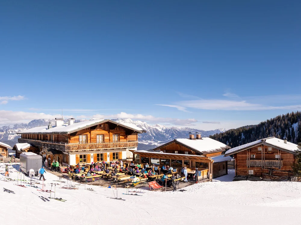 Winterurlaub beim Forstauerwirt Berghütte mit sonniger Terrasse, Gästen und Skiern im Schnee vor verschneiten Gipfeln