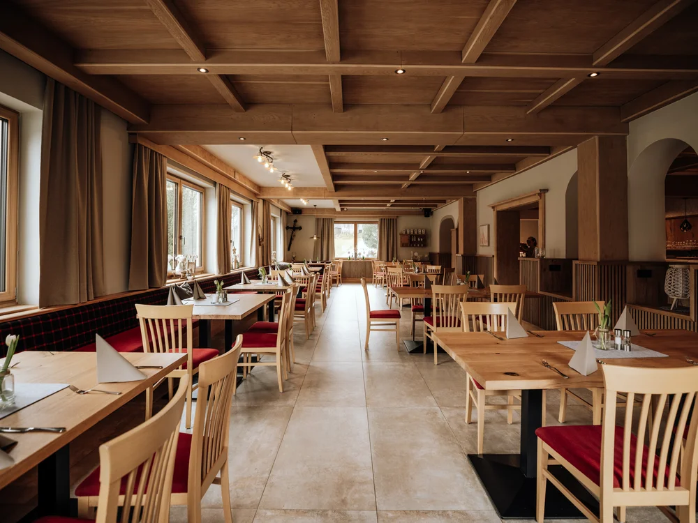 Winterurlaub beim Forstauerwirt Heller Gastraum mit Holztischen, Stühlen mit roten Sitzflächen und großen Fenstern