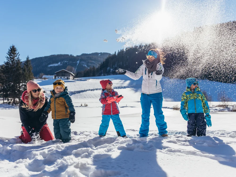 Winterurlaub beim Forstauerwirt Familie spielt im Schnee, während eine Frau eine Schneewolke aufwirbelt